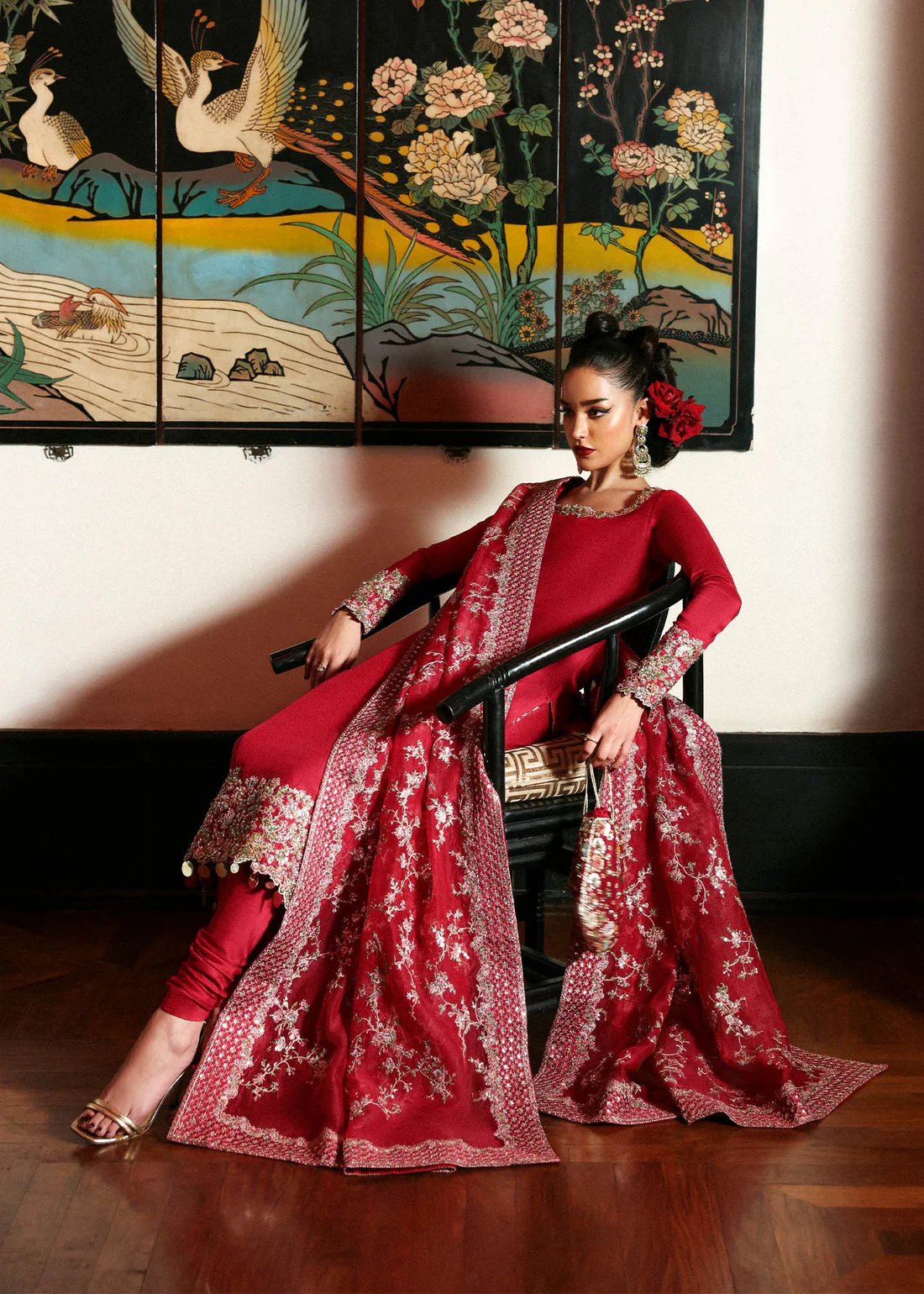 Hussain Rehar Crimson Embroidered Formal Dress The model in an embroidered deep red formal outfit seated in front of an oriental art panel.